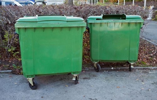 Two large green wheeled rubbish bins with closed lids positioned side by side on a paved surface, likely on a driveway or parking area in Bethnal Green, with leafless bushes and a metal fence in the background indicating an outdoor setting in late autumn or winter. The bins are made of plastic with slight surface textures, and their grey wheel assemblies are visible at the base, supporting the bins for mobility. The right bin appears slightly smaller or positioned at an angle, while the left bin is straight and firmly placed on the ground. The scene is evenly lit, with natural daylight highlighting the vibrant green colour of the bins. The setting suggests an area where Bethnal Green House Clearance services could collect household or garden waste, aligning with local rubbish removal needs. The environment is clean and typical for waste disposal in urban residential areas, with no visible debris or additional objects in the immediate vicinity.