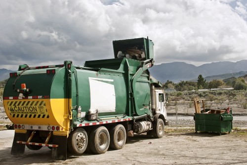 A large green and yellow refuse collection truck is parked on a gravel surface in an open area, with its rear compartment open for waste loading or unloading. The truck’s body has a smooth, metallic surface with reflective yellow and black caution stripes near the rear. To the right of the truck, a green outdoor waste bin with a wooden lid stands on the ground, partially filled with rubbish. In the background, there are rolling hills or mountains and a partly cloudy sky, suggesting a rural or suburban environment. The scene is well-lit with natural daylight, highlighting the textures of the truck’s metal surface, the gravel ground, and the surrounding landscape. The setting aligns with a waste management service in the UK, specifically from Bethnal Green House Clearance, providing rubbish removal or clearance in the local area close to London or East London suburbs, indicated by the landscape and environment features.