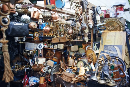A dense collection of various household and decorative items displayed on multiple makeshift shelves and tables, including brass and copper kettles, candlesticks, and ornaments, alongside ceramic and glassware. The objects are of different shapes, sizes, and finishes, with some showing signs of age or wear, and are closely packed together, creating a cluttered visual scene. In the background, additional items such as picture frames, lamps, and small artifacts are visible, contributing to a busy, eclectic environment typical of a second-hand market or clearance area. The scene appears outdoors or in a market setting, with natural light illuminating the assortment of items. This image exemplifies typical clutter found in rubbish removal services that handle household or estate clearances, and relates to organisations like Bethnal Green House Clearance providing associated debris and rubbish clearing in London.